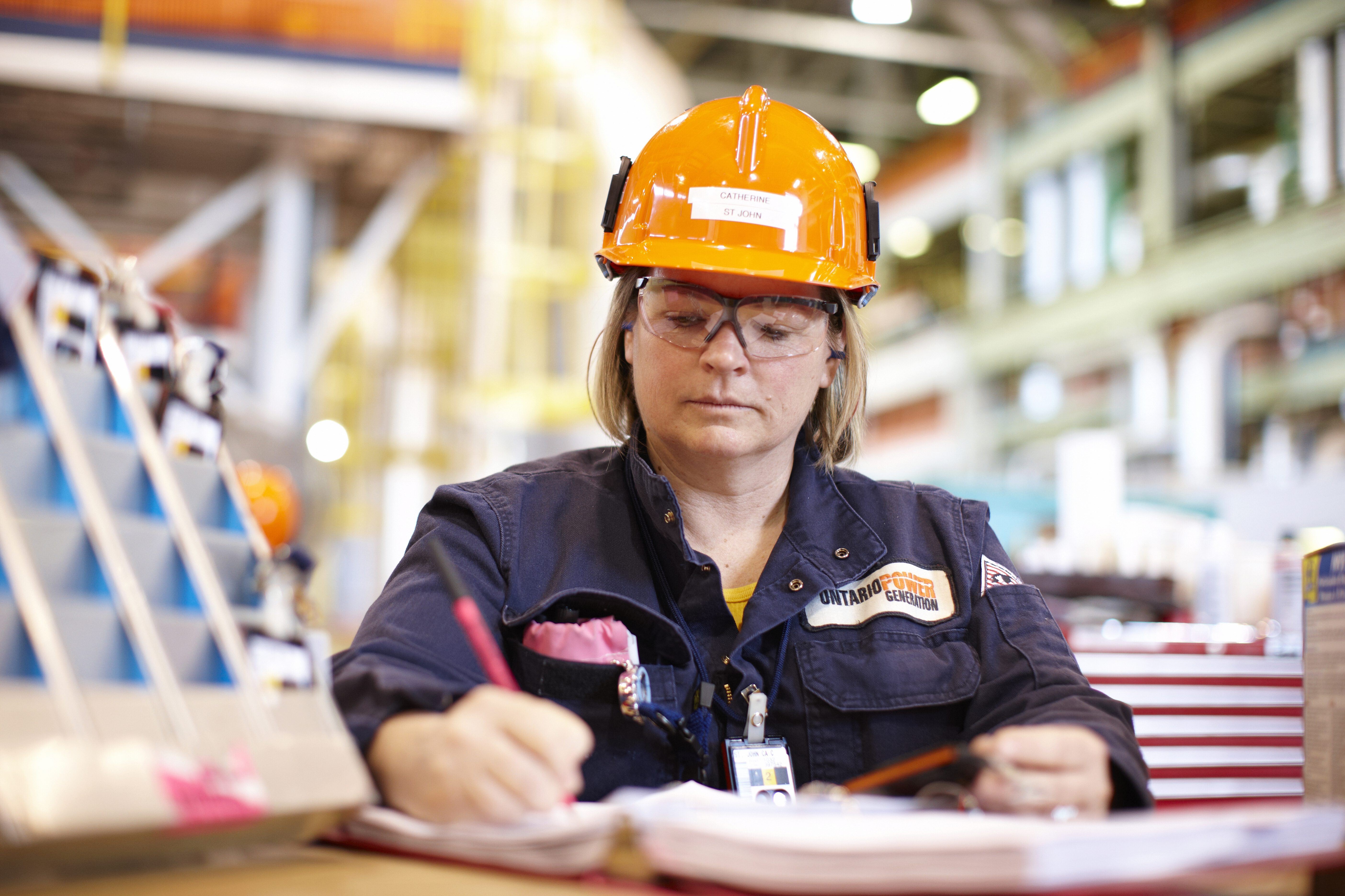 Women in OPG work overhalls and orange hardhat makes notes in a binder.