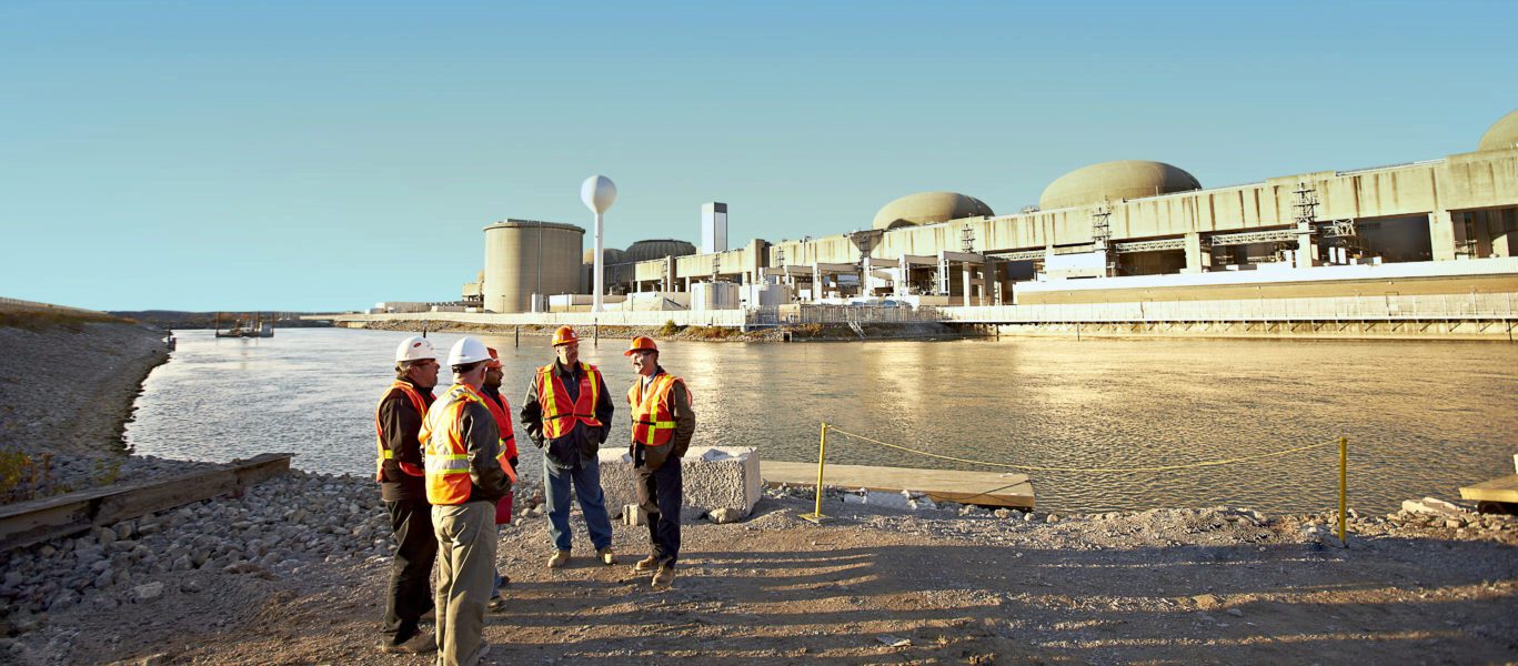 Workers in hard hats and safety vests consult across waters from Pickering Nuclear Generating Station.