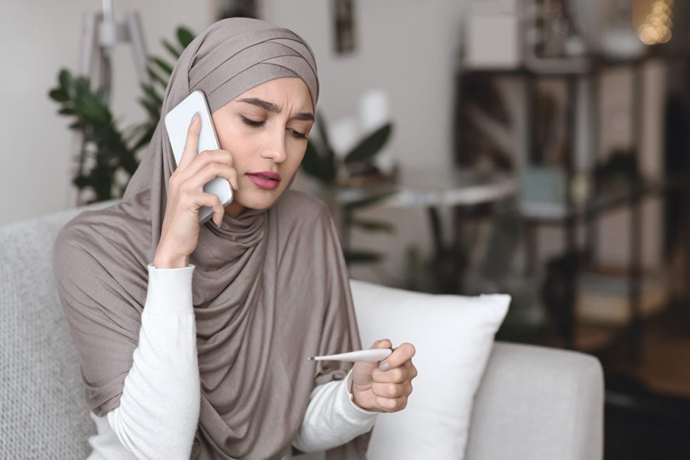 Sick arabic girl in hijab holding thermometer and calling family doctor to get consultation, sitting on couch at home