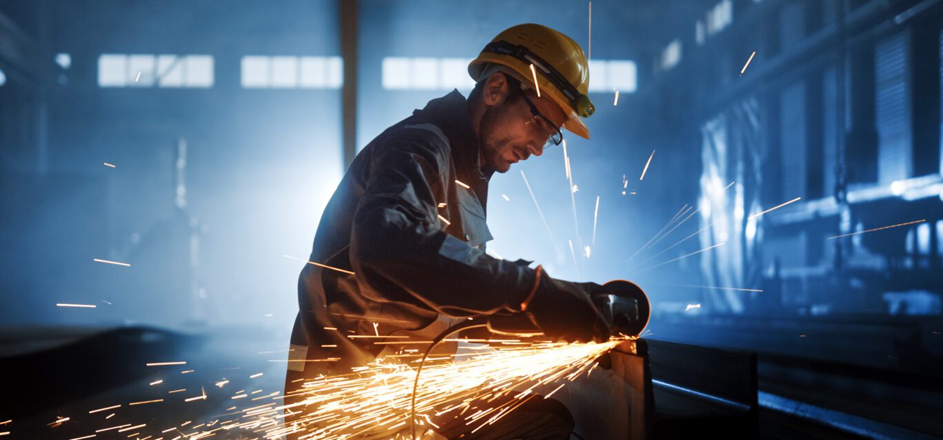 Heavy Industry Engineering Factory Interior with Industrial Worker Using Angle Grinder and Cutting a Metal Tube. Contractor in Safety Uniform and Hard Hat Manufacturing Metal Structures.