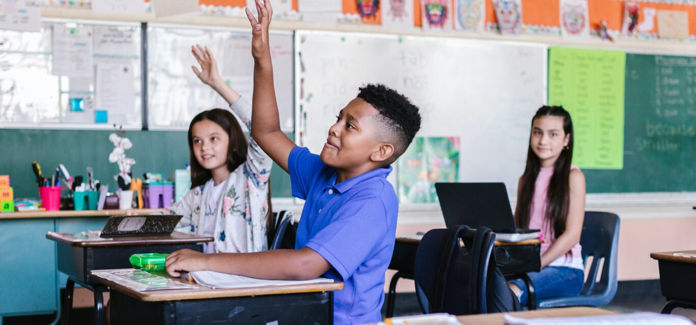 A child at a desk in a classroom raises his hand to ask a question.